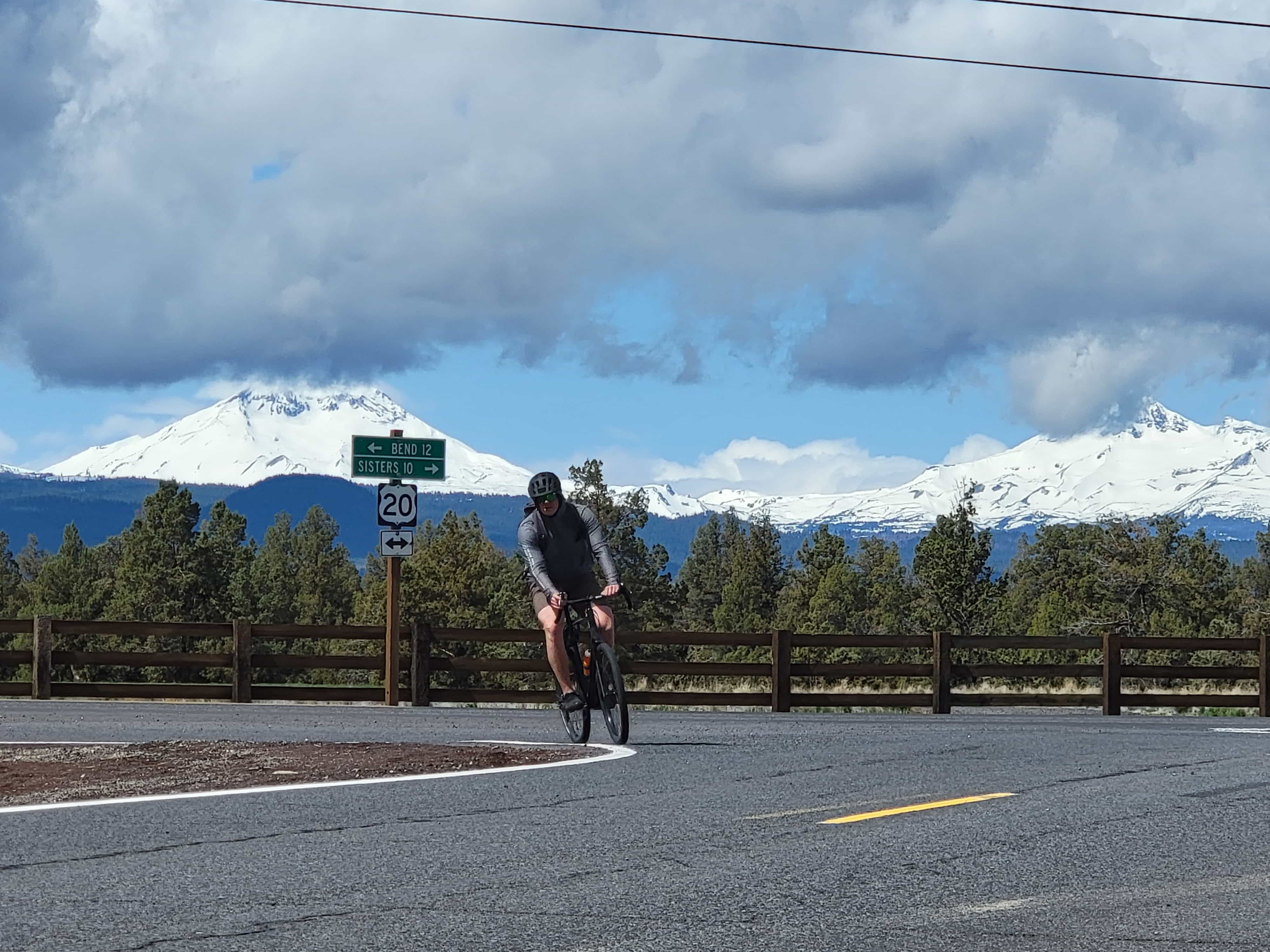 1 person taking a right hand turn on his bike towards the camera. Ranch fence and pasture in the background, with the sisters mountains behind that. A sign on the road reads "Bend 12" and "Sisters 10" in opposite directions